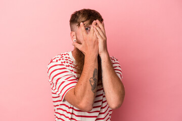Young caucasian ginger man with long beard isolated on pink background blink through fingers frightened and nervous.
