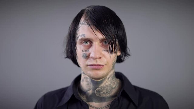 Headshot portrait of young brunette man with grey eyes and tattoos on face looking at camera. Close-up portrait of confident Caucasian guy posing at grey background with serious facial expression