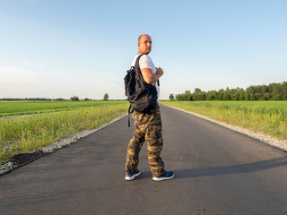 A middle-aged man with a black backpack is standing on the asphalt road, half turned around.. The...