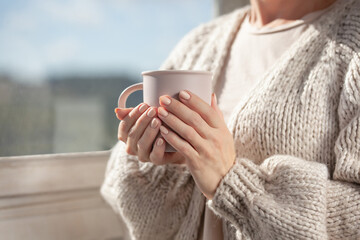 A woman in a warm cardigan by the window holds a mug in her hands. Close-up of female hands holding a cup of coffee or tea.