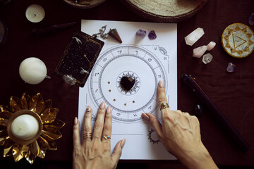 Top view of a woman pointing to a zodiac symbol in a person's birth chart on her witch altar with...