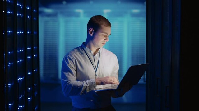 Concept of Digitalization of Information: IT Specialist In front of Server Racks with Laptop, Activates Data Center with a Touch Gesture. Working on a Laptop Standing Before Open Server Rack Cabinet