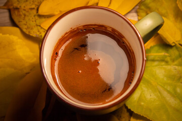 Warm cup of coffee on top of yellow leaves. Perfect moody and bright photo. Moon shape on top of the dark coffee. Autumn vibe. Green coffee mug.