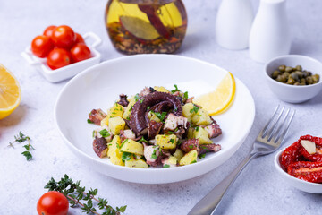 Warm salad with octopus, potatoes, tomatoes, capers and lemon on a white plate. Close-up, white background.