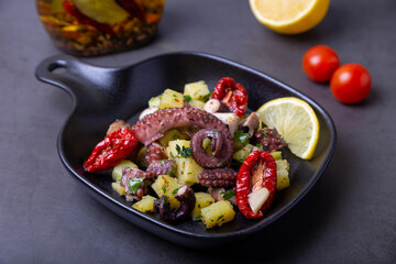 Warm salad with octopus, potatoes, sun-dried tomatoes, garlic and lemon in a black small pan. Close-up, black background.