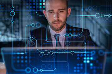 Handsome businessman in suit at workplace working with laptop to optimize development by implying new technologies in business process. Hi tech hologram over office background