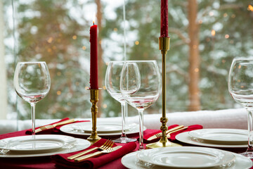 Beautiful table setting with classic white porcelain, golden cutlery and candles. Big window with winter landscape, pine tree forest covered with snow. Christmas celebration