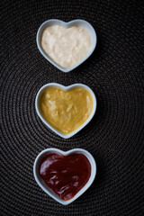 Small heart-shaped bowls with ketchup, mayonnaise and mustard sauce, vertical shot.