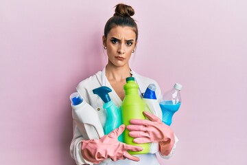 Beautiful brunette young woman wearing cleaner apron holding cleaning products clueless and confused expression. doubt concept.