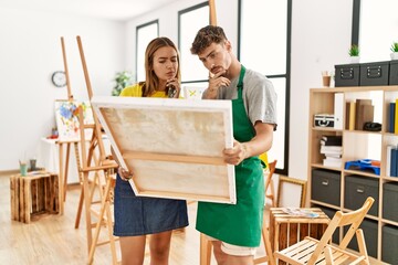 Young hispanic artist couple with serious expression holding canvas at art studio.
