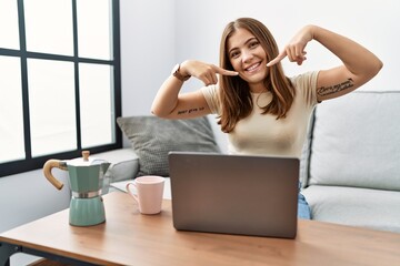 Fototapeta na wymiar Young brunette woman using laptop at home drinking a cup of coffee smiling cheerful showing and pointing with fingers teeth and mouth. dental health concept.