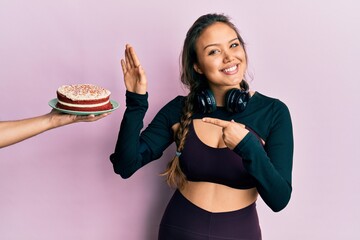 Young hispanic girl wearing sportswear rejecting cake smiling happy pointing with hand and finger