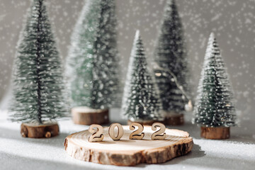 Beautiful New Year greeting card. Decorative numbers 2022 on wooden stand and green fir trees on grey blurred background with snow. Happy new year 2022.