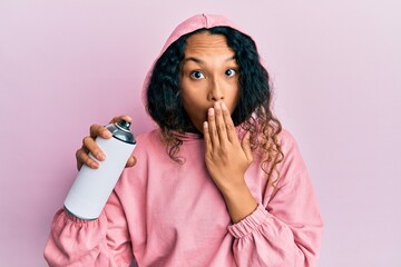 Young latin woman wearing sweatshirt holding graffiti spray covering mouth with hand, shocked and...