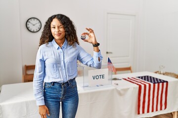 Beautiful hispanic woman standing by at political campaign by voting ballot winking looking at the...