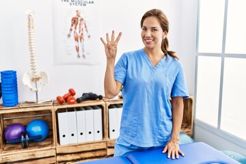 Middle age hispanic physiotherapist woman working at pain recovery clinic showing and pointing up with fingers number four while smiling confident and happy.