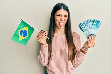 Young hispanic woman holding brazil flag and real banknotes sticking tongue out happy with funny...