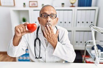 Mature doctor man holding red heart at the clinic covering mouth with hand, shocked and afraid for mistake. surprised expression