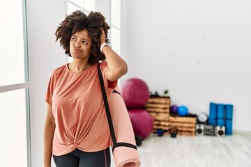 Plakat African american woman with afro hair holding yoga mat at pilates room confuse and wondering about question. uncertain with doubt, thinking with hand on head. pensive concept.