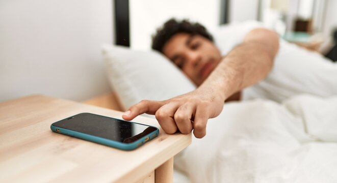 Young hispanic man turning off smartphone alarm lying on the bed at bedroom.