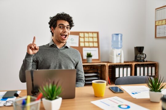 Young hispanic man wearing business style sitting on desk at office pointing finger up with successful idea. exited and happy. number one.