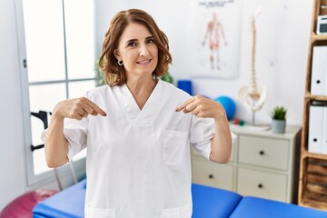Middle age physiotherapist woman working at pain recovery clinic looking confident with smile on face, pointing oneself with fingers proud and happy.