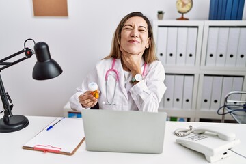 Young hispanic woman wearing doctor uniform holding pills at the clinic touching painful neck, sore throat for flu, clod and infection
