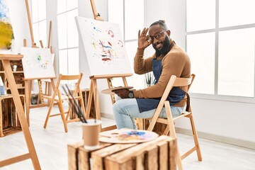 African american artist man painting on canvas at art studio smiling happy doing ok sign with hand on eye looking through fingers