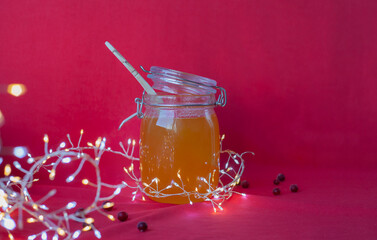 Big jar of honey with wooden spoon inside on red background with Christmas decoration. Natural nutrition during winter holidays. Winter poster or banner with organic honey, cranberry and string lights