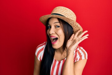 Young latin woman wearing summer hat smiling with hand over ear listening and hearing to rumor or gossip. deafness concept.