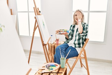 Young artist woman painting on canvas at art studio covering one eye with hand, confident smile on face and surprise emotion.