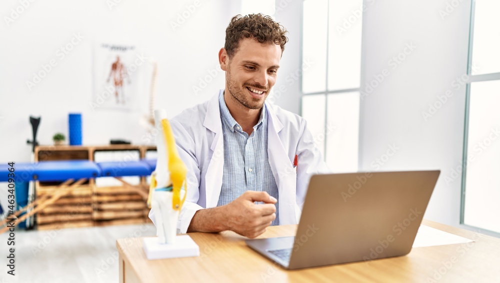 Wall mural Young hispanic man wearing physiotherapist uniform having video call working at clinic