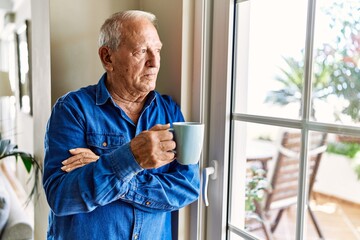 Senior man with grey hair leaning by the window of his home, drinking a cup of coffee in the morning