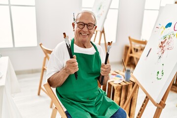 Senior grey-haired artist man smiling happy holding paintbrushes at art studio.