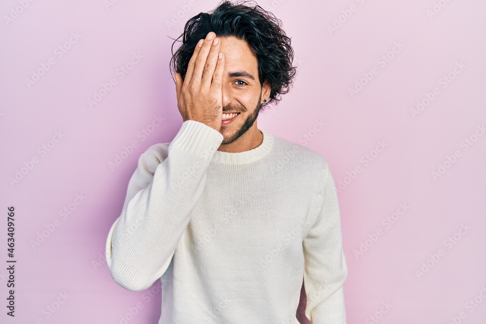 Sticker Handsome hispanic man wearing casual white sweater covering one eye with hand, confident smile on face and surprise emotion.