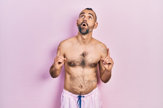 Young hispanic man shirtless wearing swimsuit amazed and surprised looking up and pointing with fingers and raised arms.