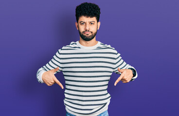 Young arab man with beard wearing casual striped sweater pointing down looking sad and upset, indicating direction with fingers, unhappy and depressed.