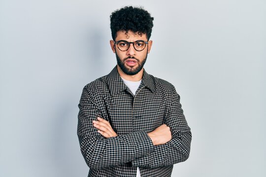 Young arab man with beard wearing glasses with arms crossed gesture in shock face, looking skeptical and sarcastic, surprised with open mouth