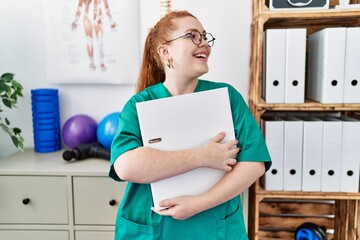 Young redhead woman wearing phsiologist uniform holding binder at clinic