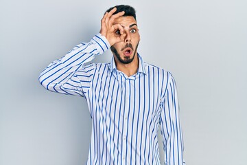 Young hispanic man with beard wearing casual striped shirt doing ok gesture shocked with surprised face, eye looking through fingers. unbelieving expression.