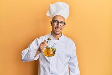 Bald man with beard wearing professional cook apron holding olive oil looking positive and happy standing and smiling with a confident smile showing teeth