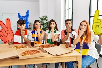 Group of young hispanic people eating pizza supporting soccer team at home covering mouth with hand, shocked and afraid for mistake. surprised expression