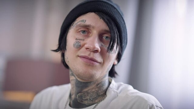 Close-up tattooed face of young man in hat looking at camera smiling. Headshot portrait of confident relaxed millennial Caucasian guy posing indoors at home. Lifestyle and style concept