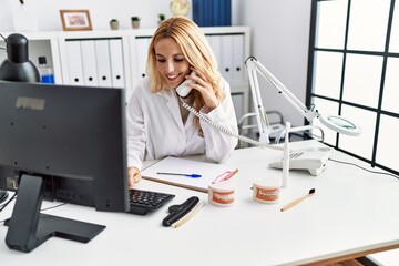 Young blonde woman wearing dentist uniform speaking on the phone at dental clinic - Powered by Adobe