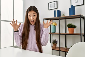 Young chinese girl wearing casual clothes sitting on the table at home celebrating victory with happy smile and winner expression with raised hands