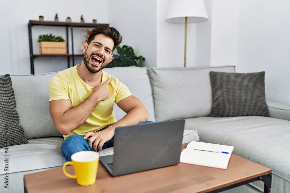 Poster Young man with beard using laptop at home cheerful with a smile of face pointing with hand and finger up to the side with happy and natural expression on face