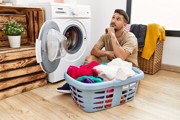 Young handsome man putting dirty laundry into washing machine with hand on chin thinking about question, pensive expression. smiling with thoughtful face. doubt concept.