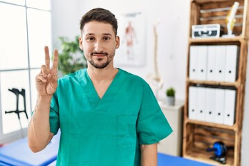 Young physiotherapist man working at pain recovery clinic showing and pointing up with fingers number two while smiling confident and happy.