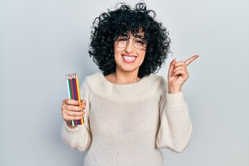 Young middle east woman holding colored pencils smiling happy pointing with hand and finger to the side
