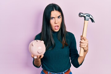 Young brunette woman holding piggy bank and hammer in shock face, looking skeptical and sarcastic, surprised with open mouth